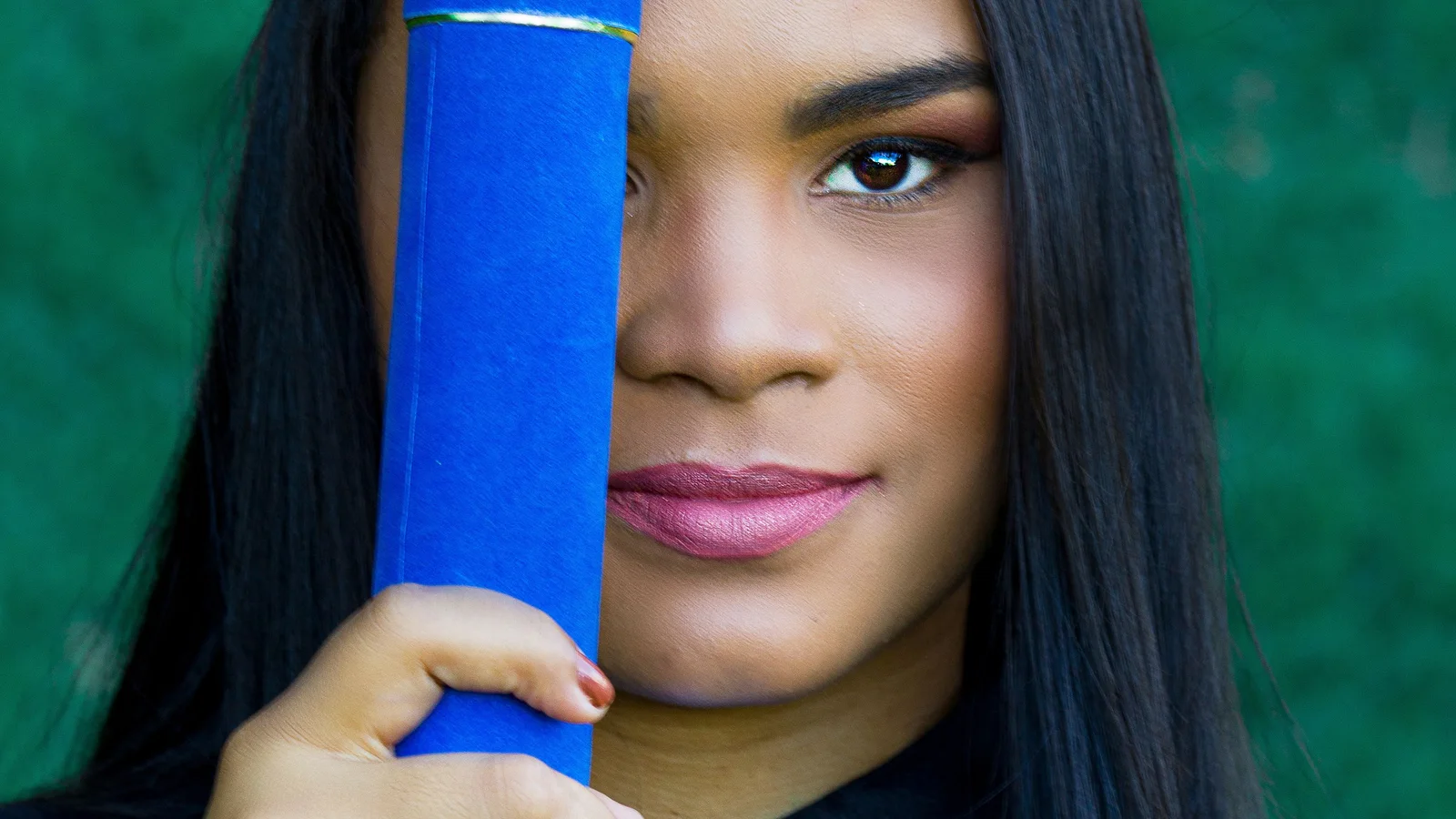 A woman in a graduation cap holding a rolled blue document beside her face.