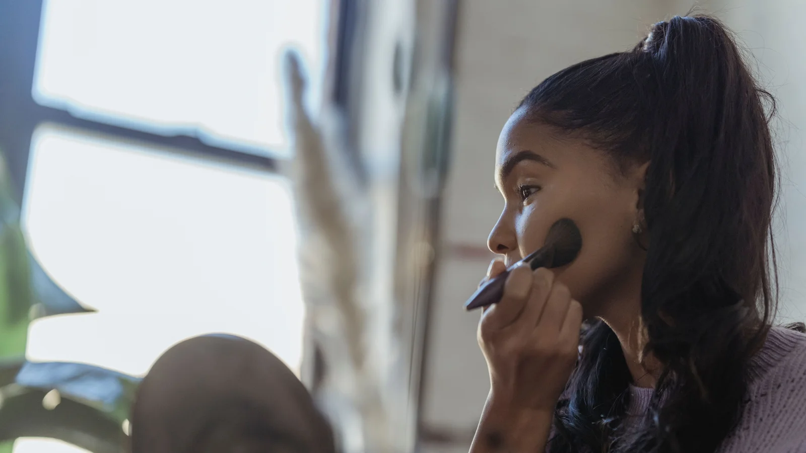 A woman applying make-up to her cheek with a large brush beside a mirror.