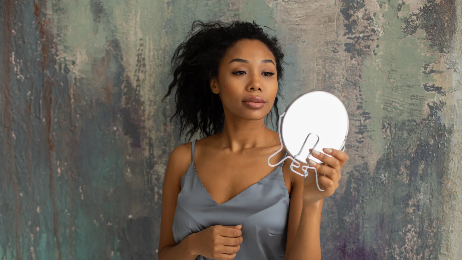 A woman holding a small round mirror and looking at her reflection.