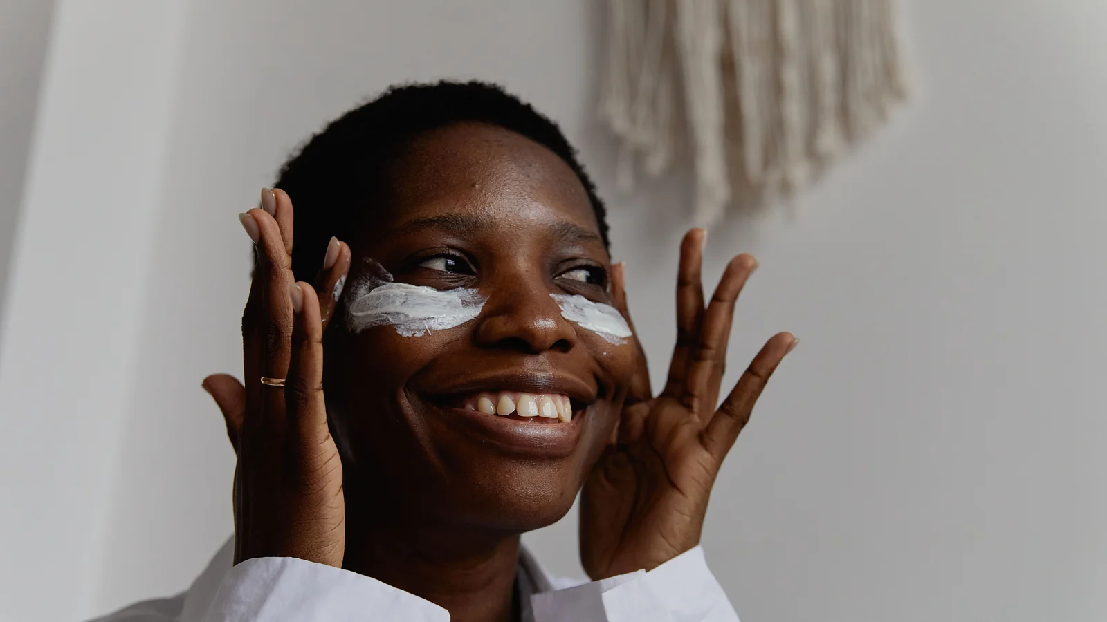A smiling woman applying white cream beneath her eyes.