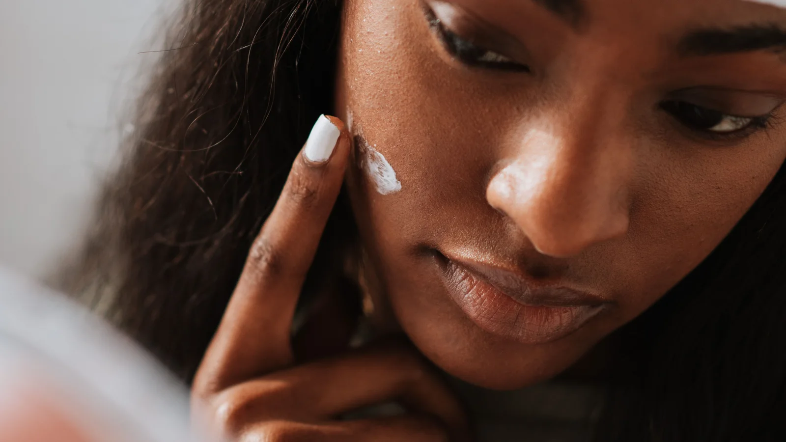 A woman in a white headband placing cream on her cheek beside a mirror.