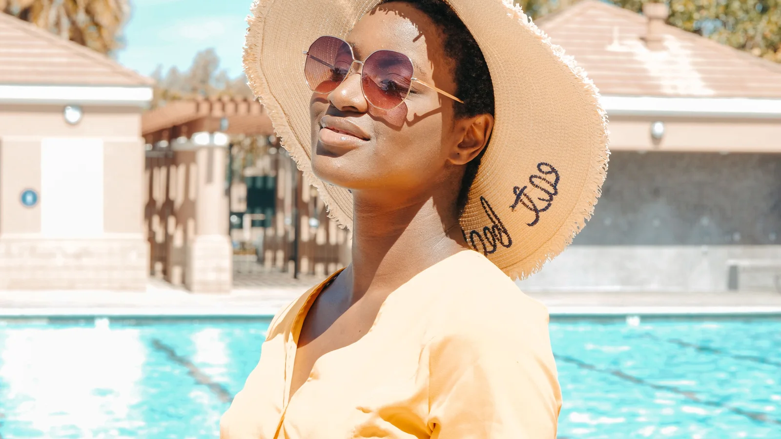 A woman wearing a broad-brimmed hat, sunglasses and a pale shirt beside a pool.