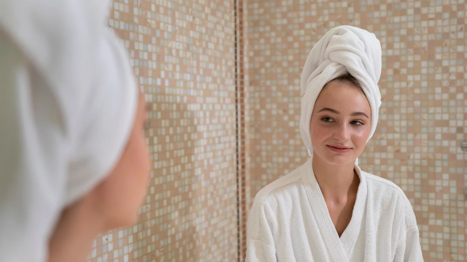 A woman wearing a white robe and towel looking at her reflection.