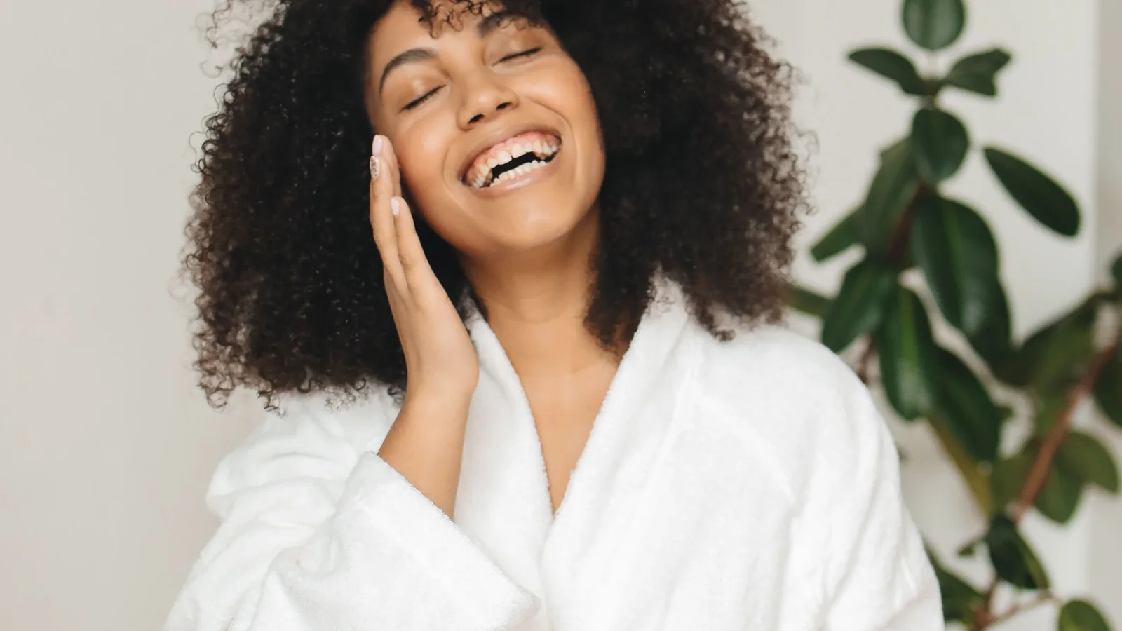 A smiling woman in a white robe touching cream to her cheek.