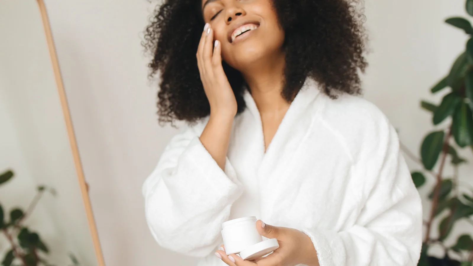 A smiling woman in a white robe applying cream from a small jar.