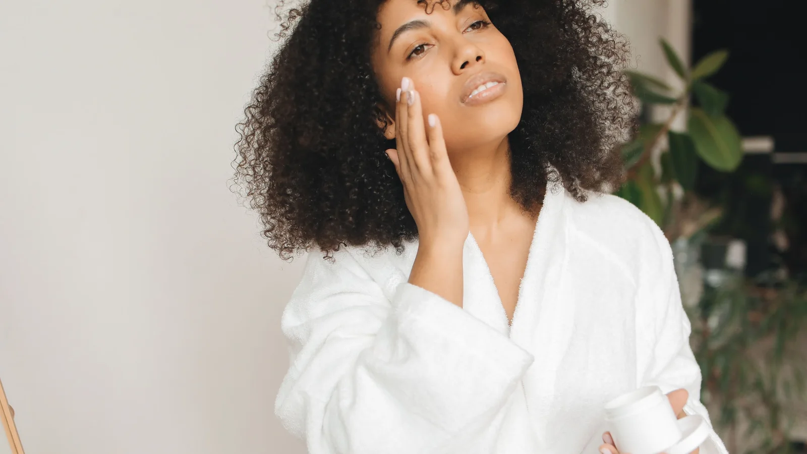 A woman in a white robe touching cream to her cheek beside a green plant.