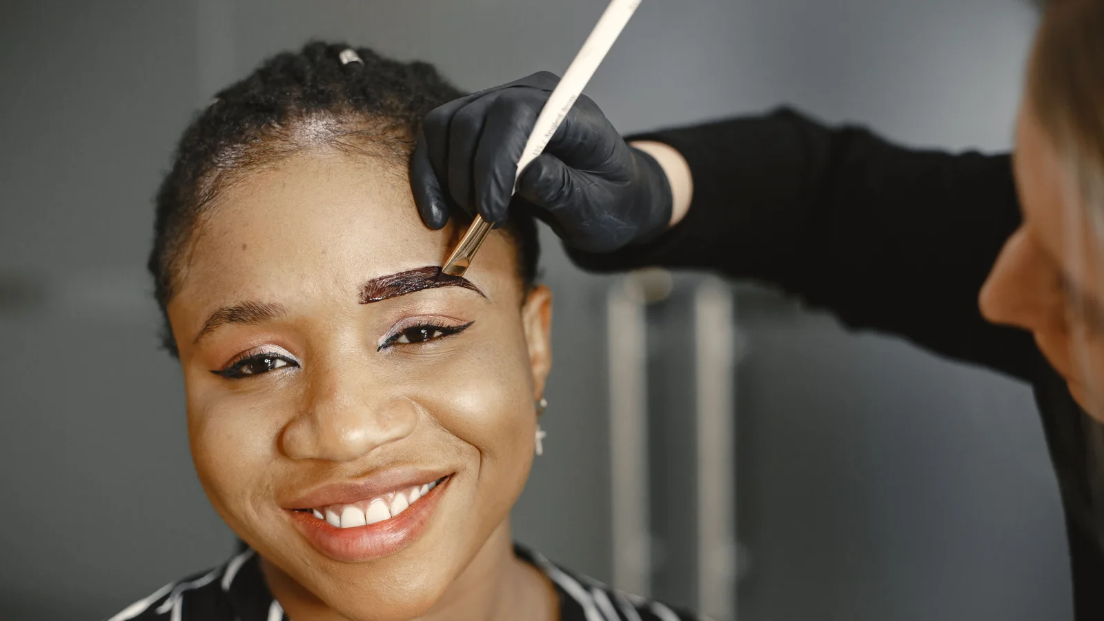 A gloved practitioner grooming a smiling woman's eyebrow with a small tool.