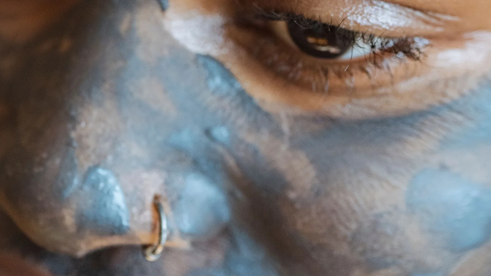 Close-up of a woman's face with a grey cosmetic mask and a nose ring.