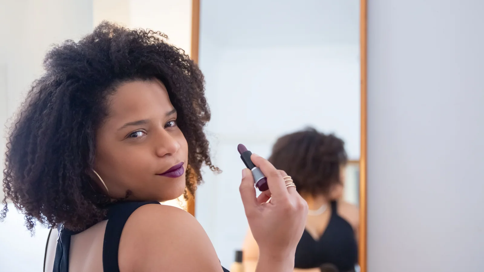 A woman holding lipstick near her lips while turning towards a mirror.