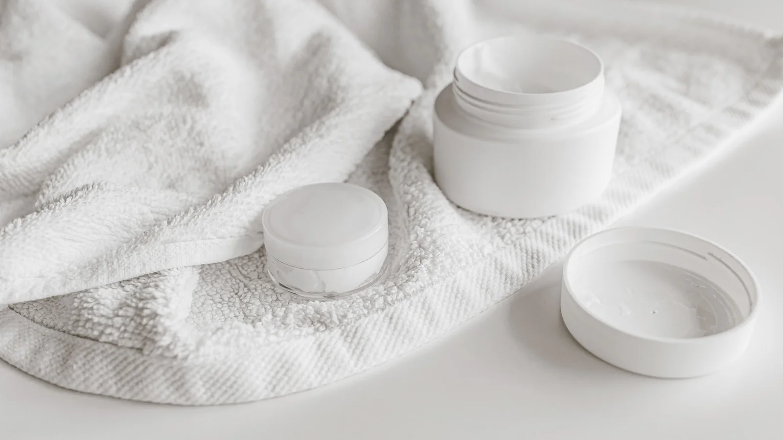An open white cosmetic jar and its lid on a folded towel.