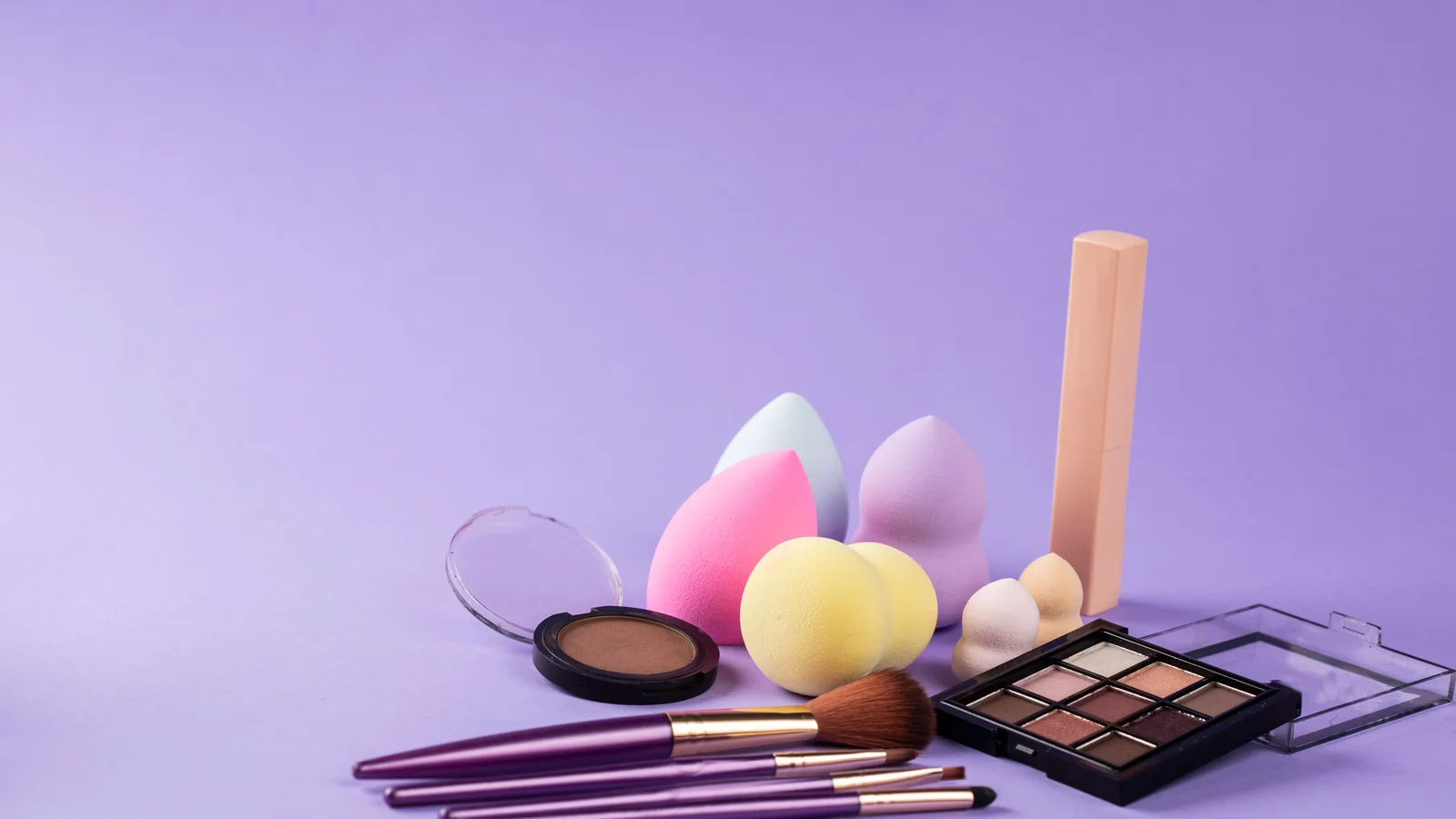 Make-up sponges, brushes and palettes arranged on a purple surface.