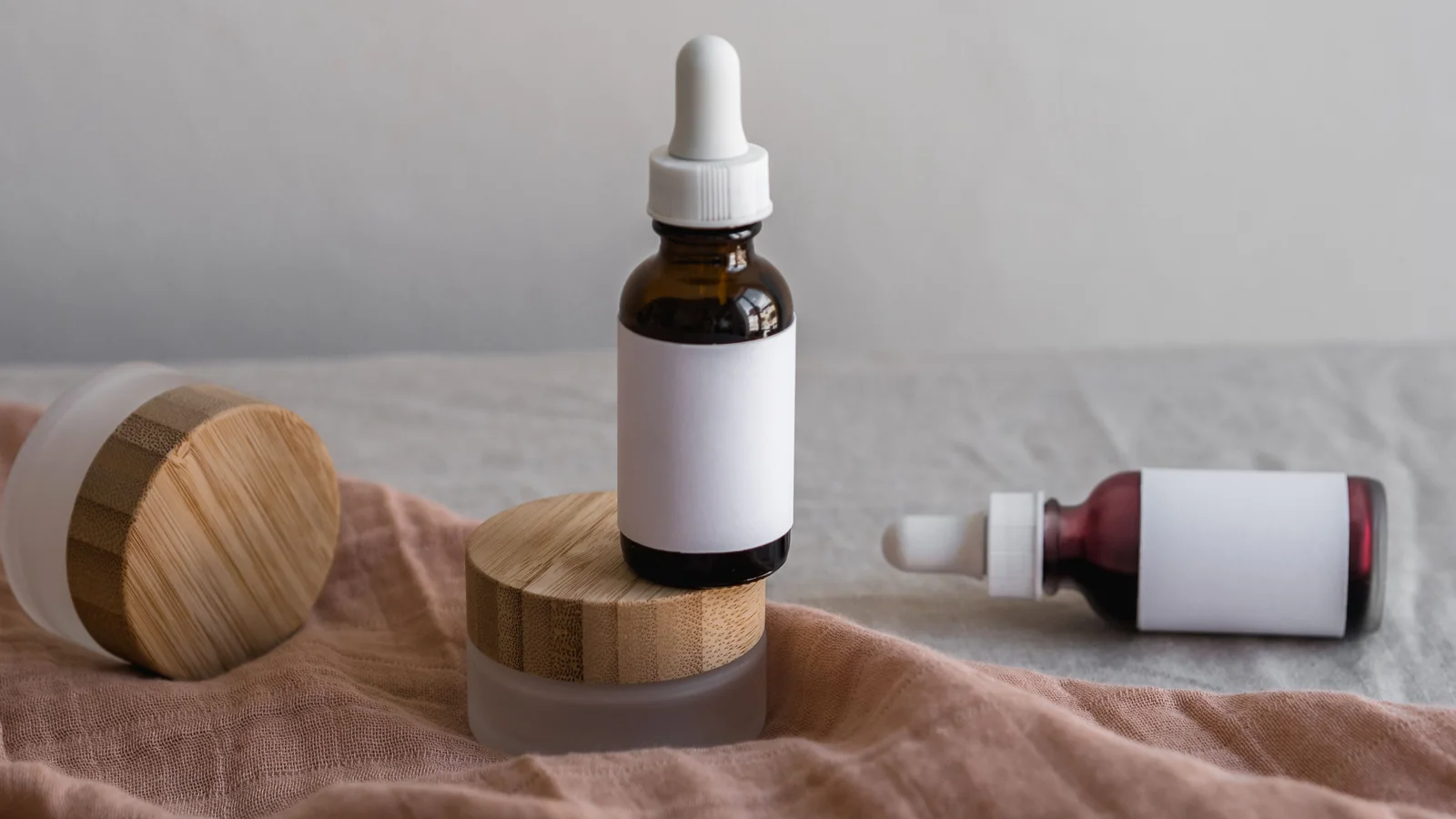 Cosmetic dropper bottles and a wooden-lidded jar on pale cloth.