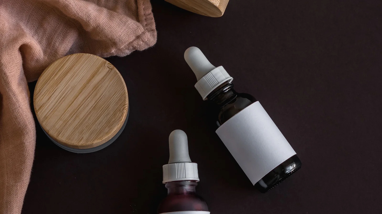 Cosmetic bottles and jars with pale labels and wooden lids on dark fabric.