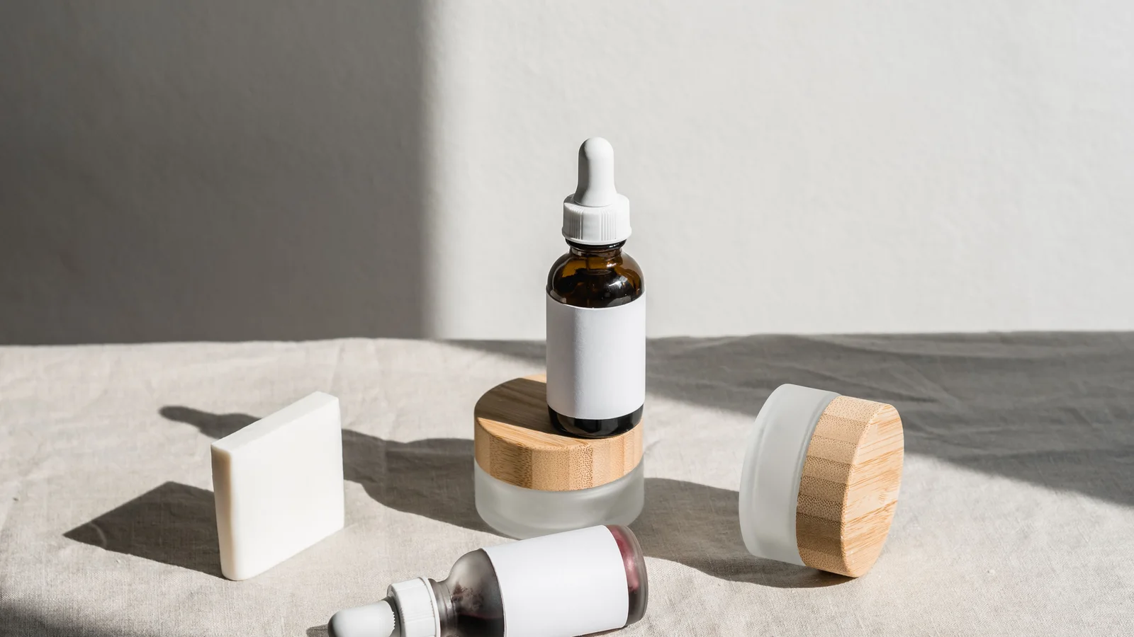 A cosmetic bottle, jar and bar arranged on grey fabric in sunlight.