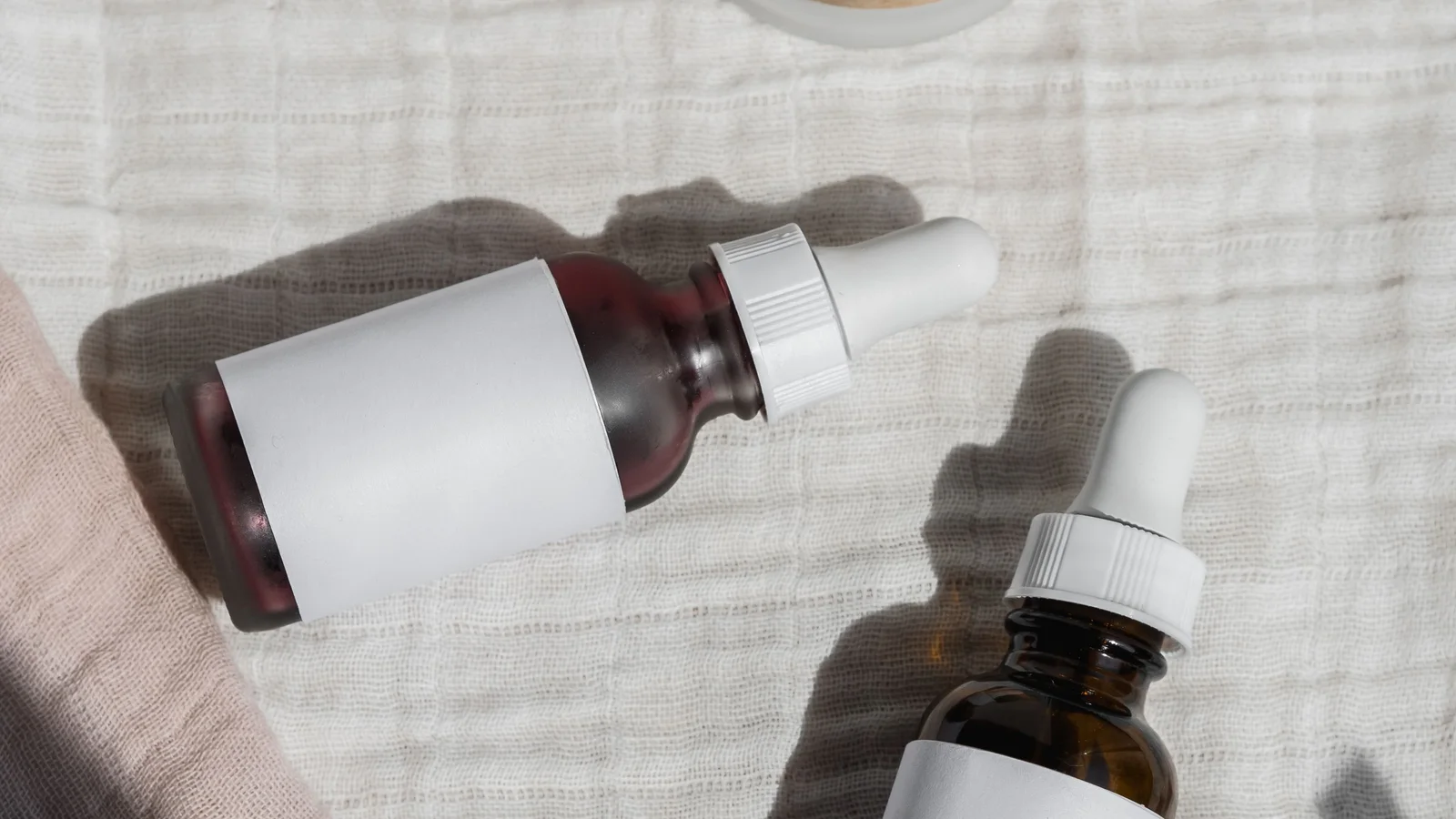 Two cosmetic dropper bottles and a wooden jar lid on textured fabric.