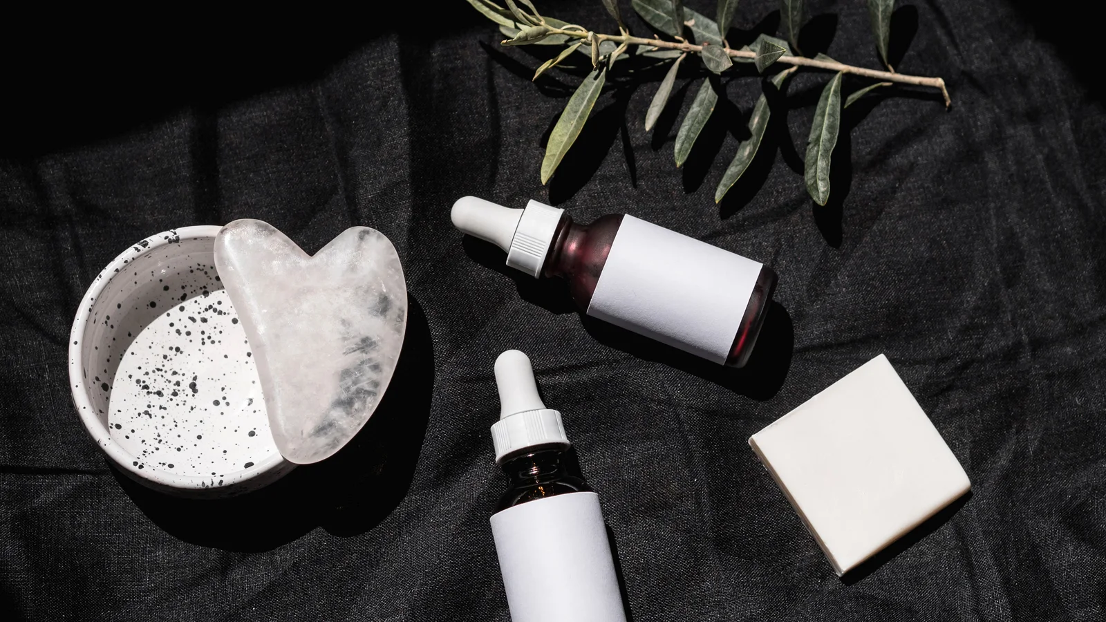 Cosmetic bottles, a pale facial tool and leaves arranged on dark fabric.