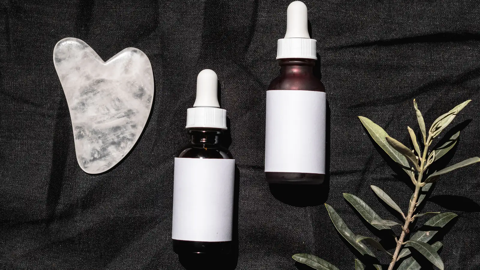 Two cosmetic dropper bottles, a pale facial tool and leaves on dark fabric.