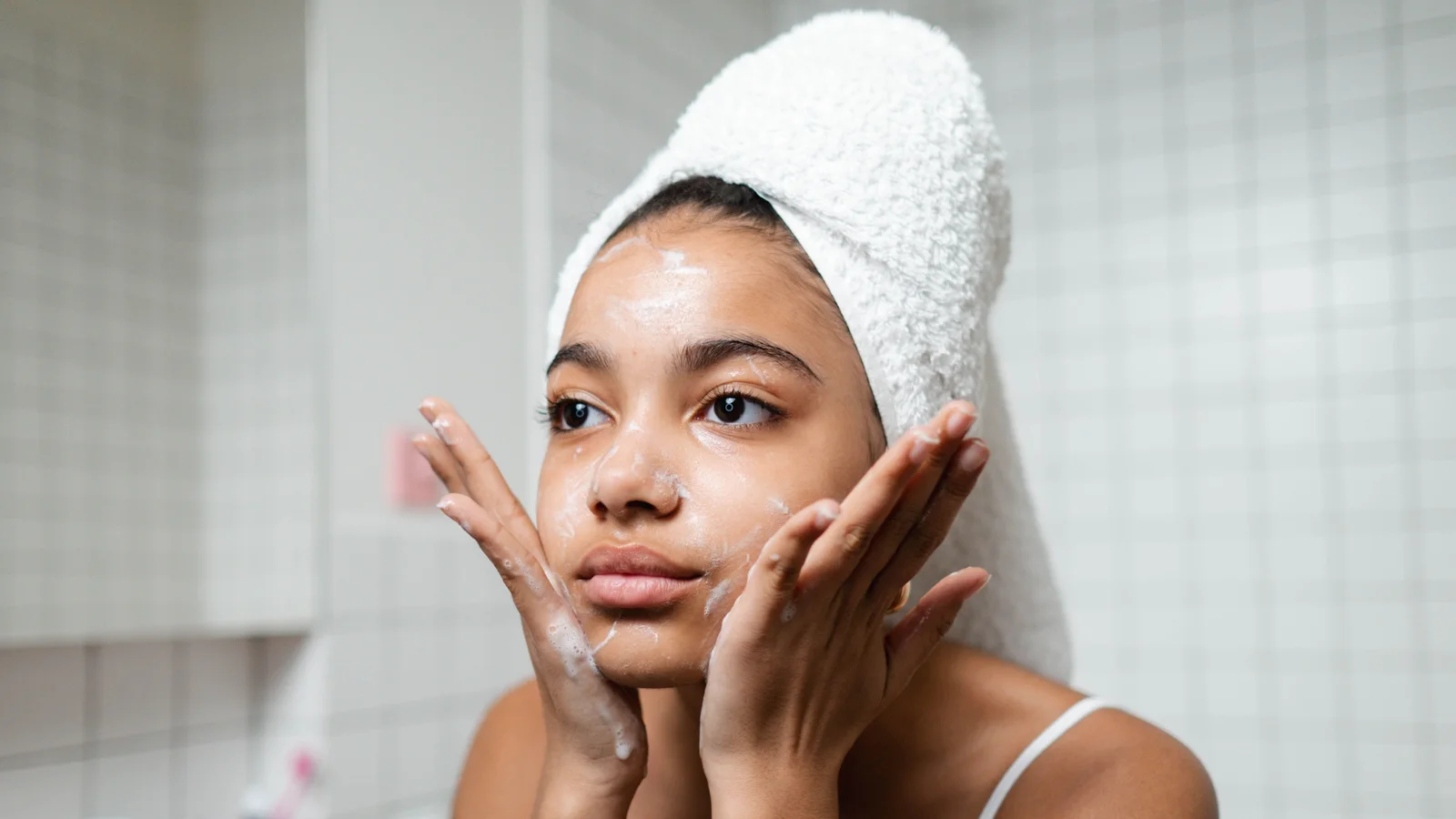 A woman gently touching both cheeks with cream visible on her face.