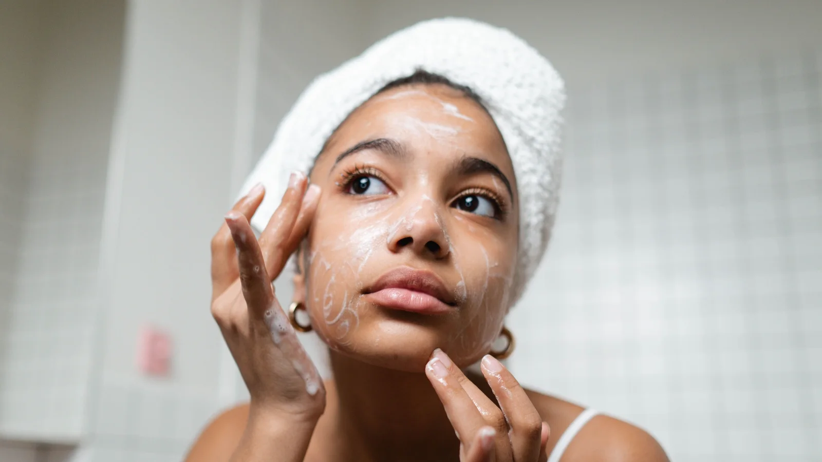 A woman touching a small amount of cream to her face beside a mirror.