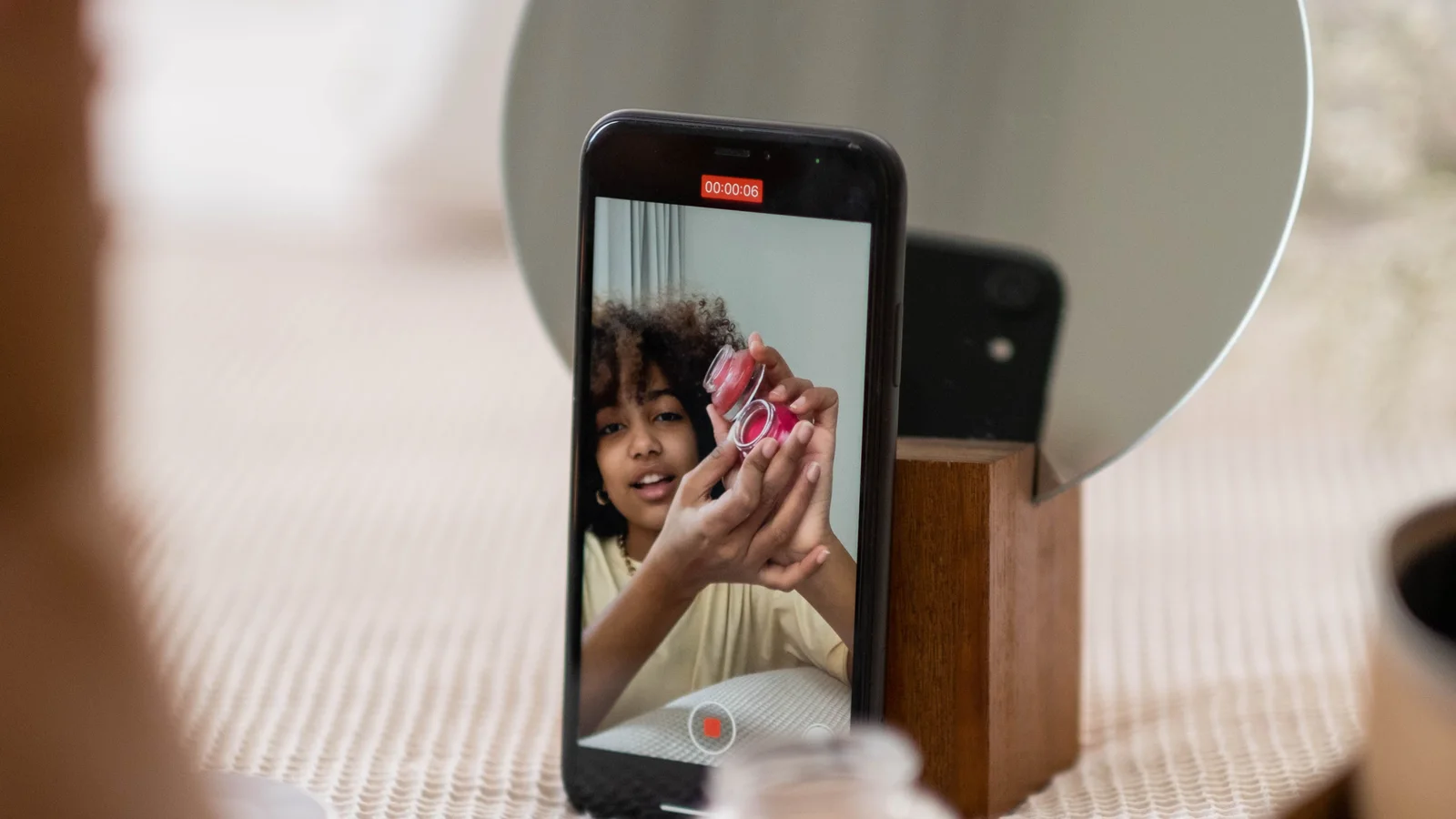 A woman filming with a phone beside cosmetics and a round mirror.