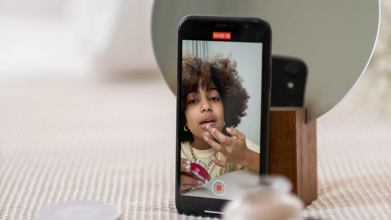 A phone on a stand recording a woman seated at a table with beauty products.