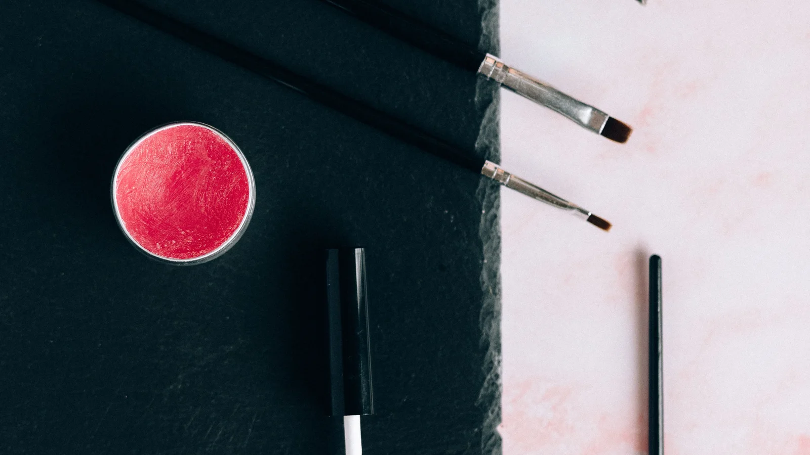 Cosmetic pencils, brushes, a pot and a gloss tube arranged on a black tray.