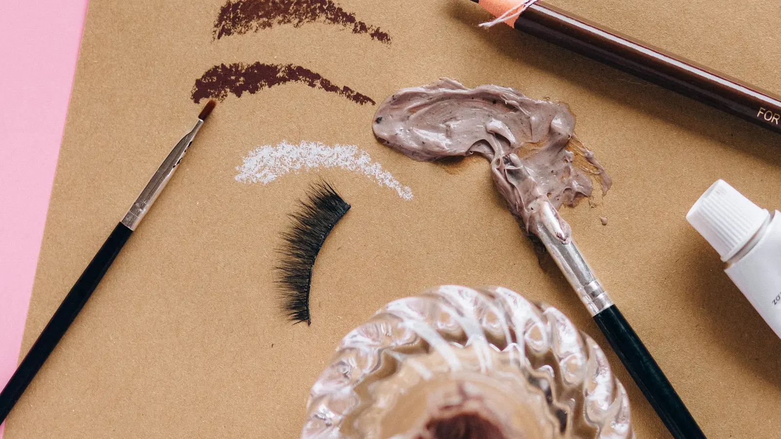 A cosmetic pencil and make-up textures arranged on pink and brown paper.
