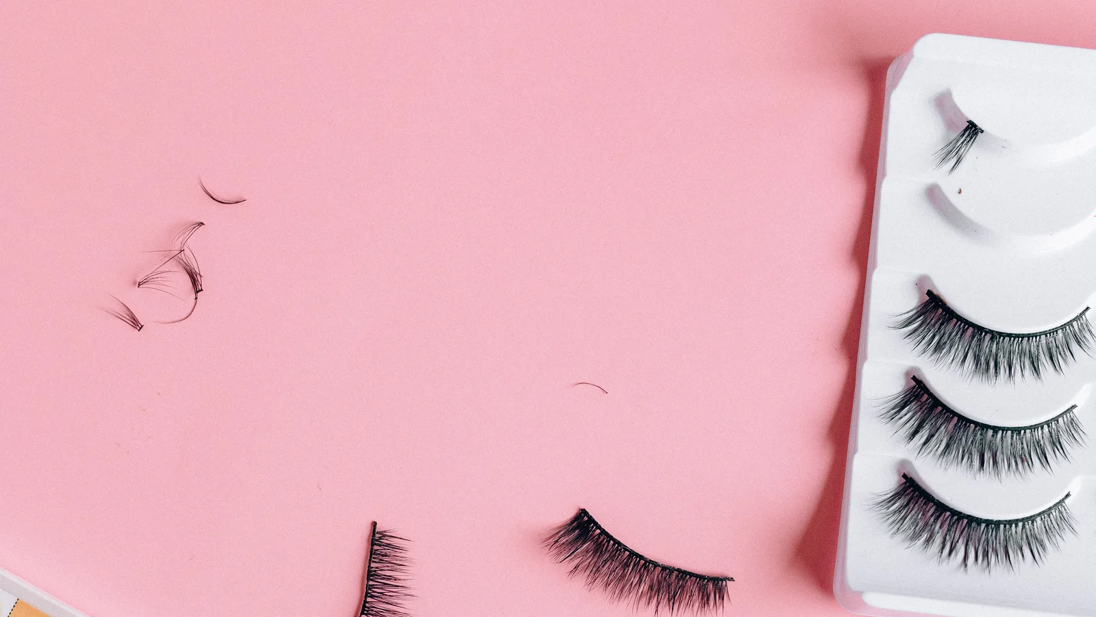 False eyelashes, a small brush and cosmetics arranged on a pink surface.