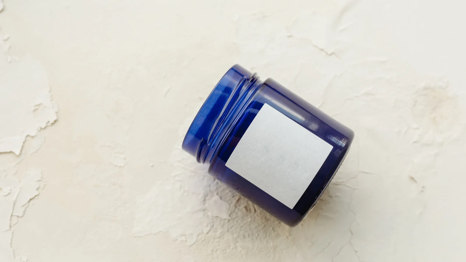 An open blue cosmetic jar with a blank white label on a pale surface.