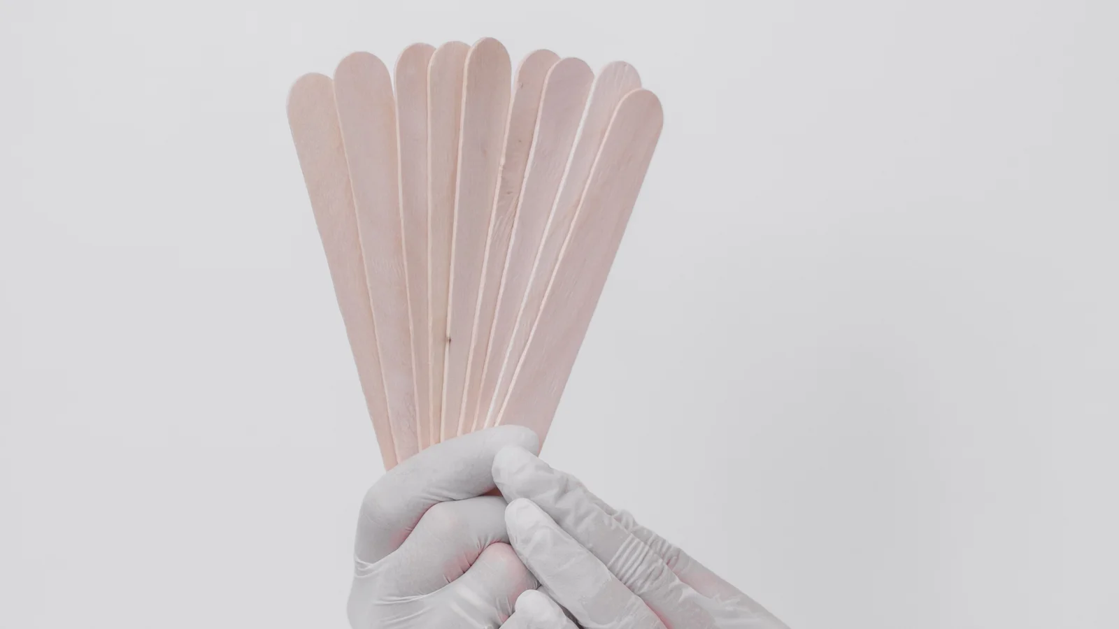 A gloved hand holding several flat wooden spatulas against a pale background.