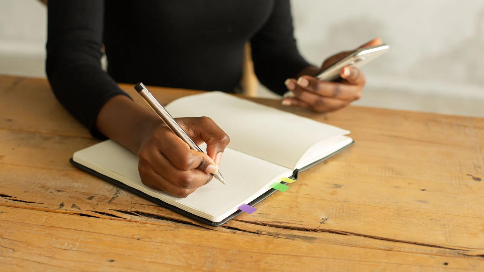 A woman writing in a notebook while holding a phone at a wooden table.