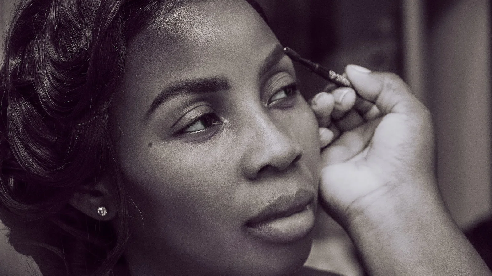 Black-and-white portrait of a woman having make-up applied near one brow.