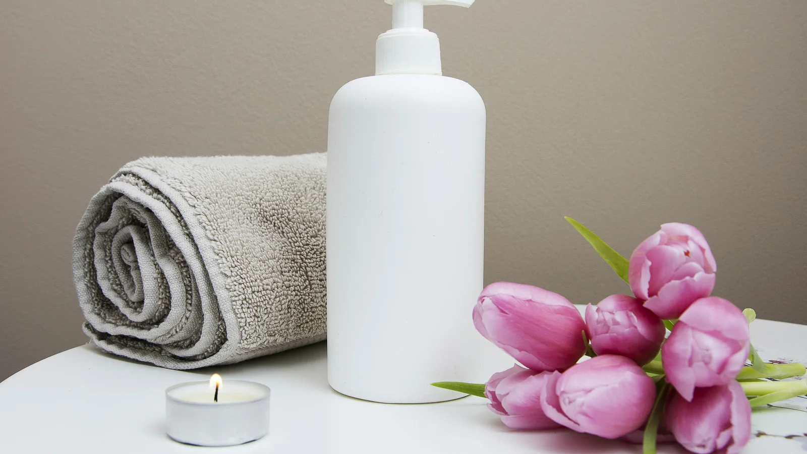 A white pump bottle beside a folded towel and pink flowers.